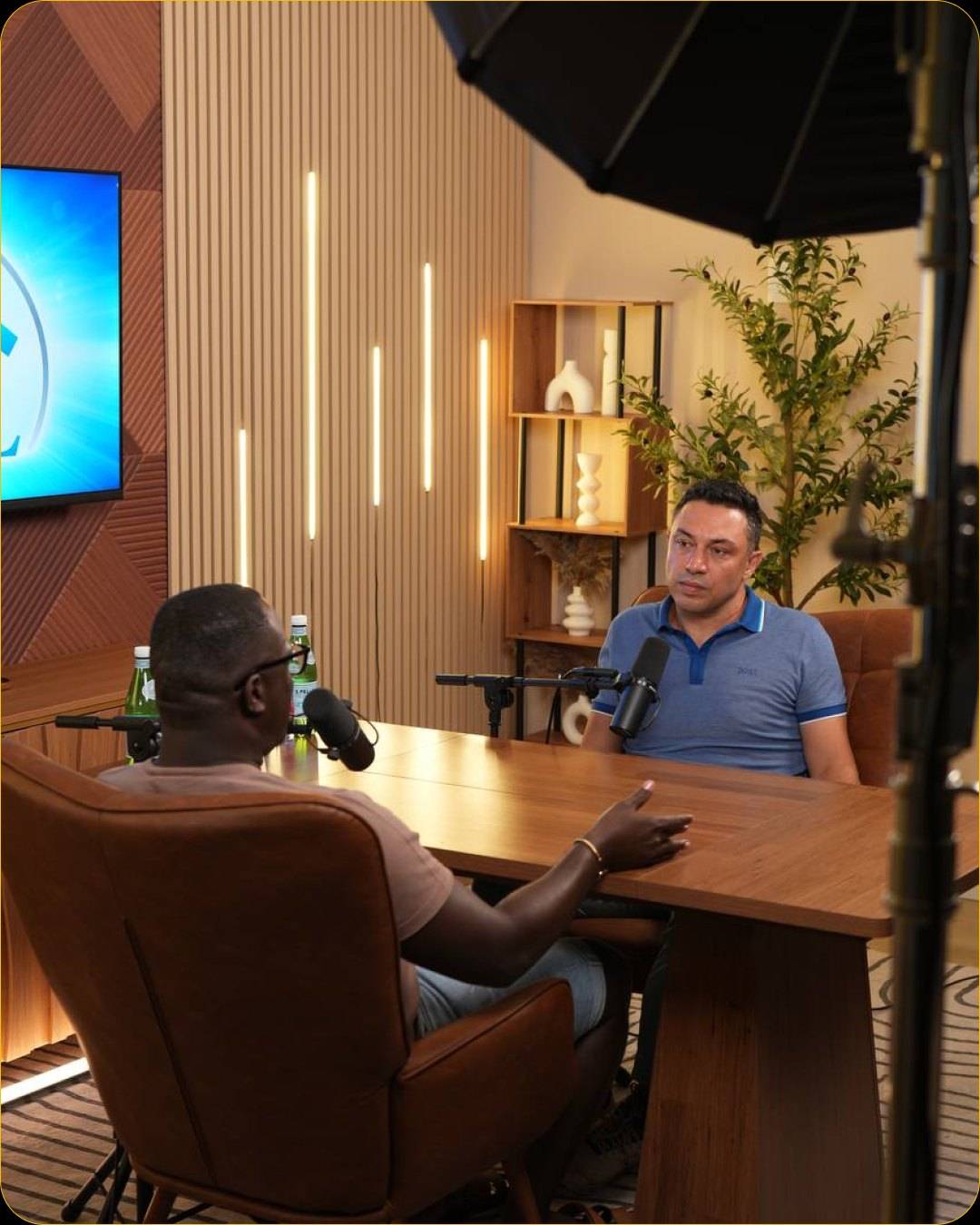 Studio #4 at The Cast Studio: Two people across the wooden table, each with a microphone on a boom arm, with the wall display and LED light bars behind them.