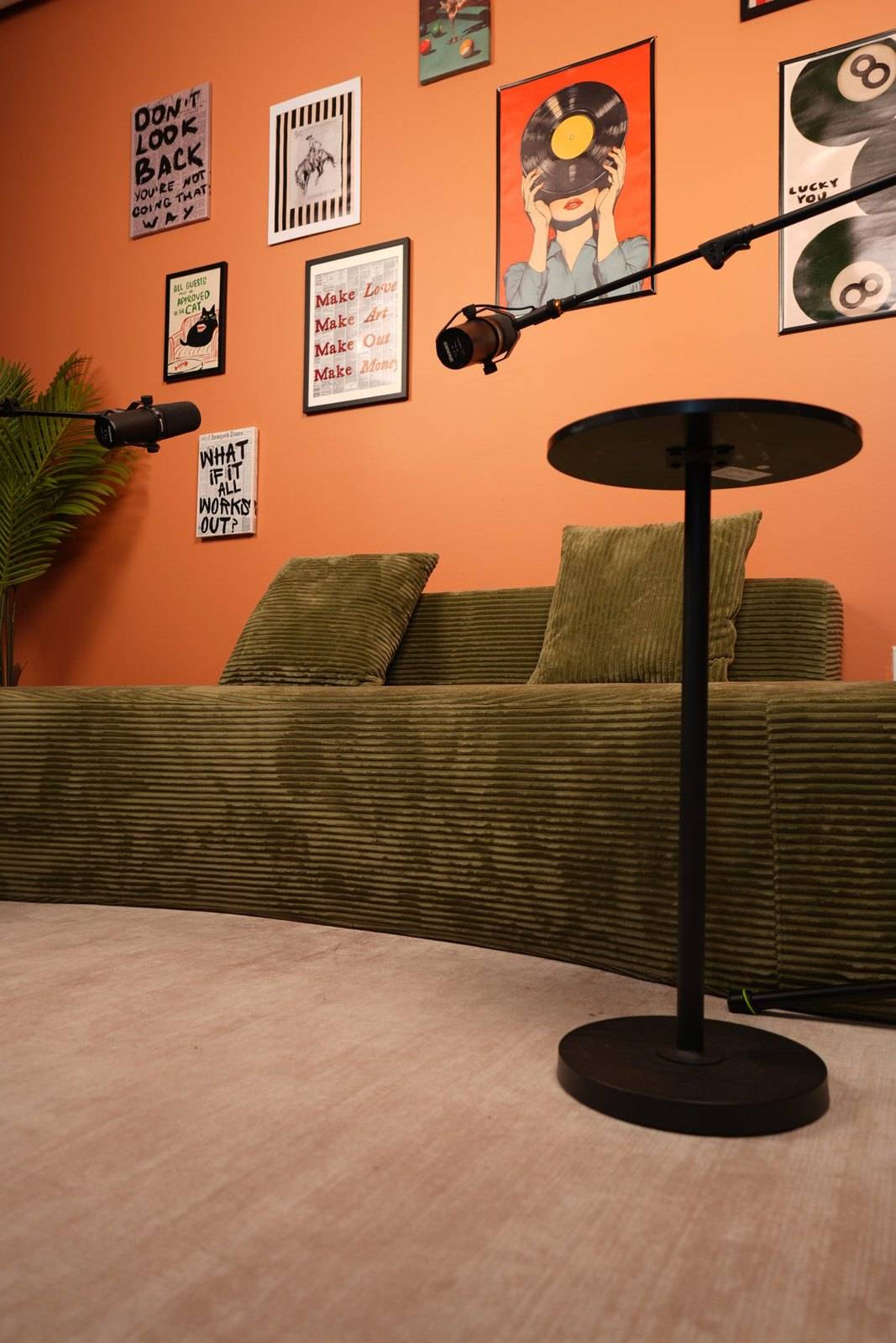 Studio #6 at The Cast Studio: A green corduroy sofa against the orange gallery wall, with two microphones on boom arms and a small side table in front.