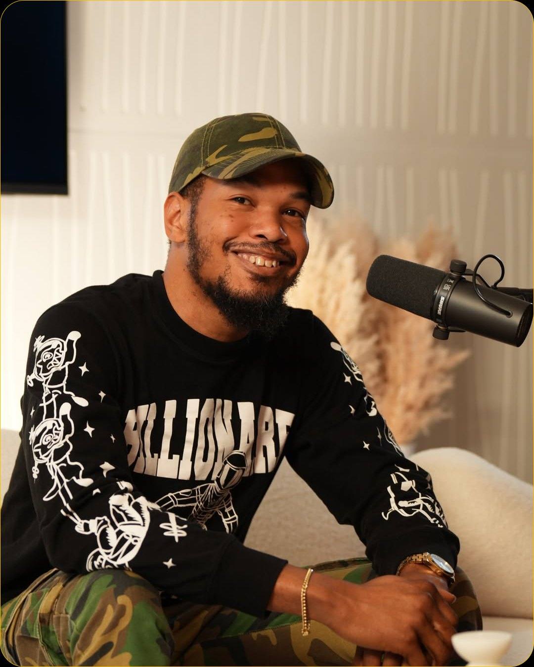 A smiling guest in a camouflage cap at the microphone on the white sofa of Studio #2