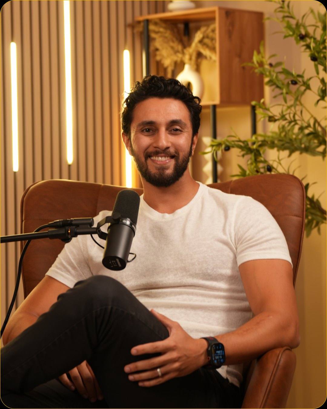 A smiling guest in a white T-shirt at the microphone in a leather armchair in Studio #4, with LED light bars and a plant behind him